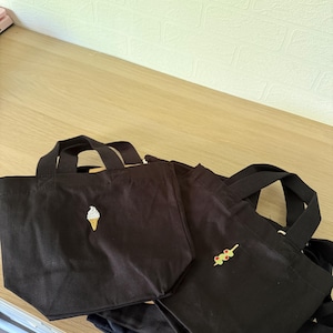 K&ouml;nnte beinhalten: Zwei schwarze Tragetaschen aus Segeltuch mit kurzen Henkeln. Die linke Tasche ist mit einer kleinen wei&szlig;en Eiswaffel bestickt. Die rechte Tasche zeigt eine kleine Stickerei eines gr&uuml;nen Oliven-Spie&szlig;es mit roten Pimentost&uuml;cken.