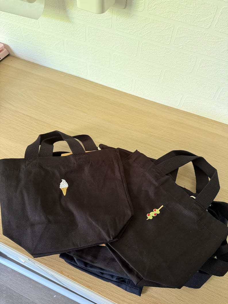 K&ouml;nnte beinhalten: Zwei schwarze Tragetaschen aus Segeltuch mit kurzen Henkeln. Die linke Tasche ist mit einer kleinen wei&szlig;en Eiswaffel bestickt. Die rechte Tasche zeigt eine kleine Stickerei eines gr&uuml;nen Oliven-Spie&szlig;es mit roten Pimentost&uuml;cken.
