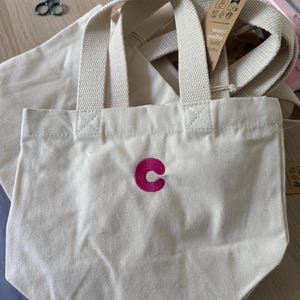 K&ouml;nnte beinhalten: Eine kleine, cremewei&szlig;e Canvas-Tragetasche mit zwei stabilen Henkeln und einem leuchtend pinkfarbenen, gestickten Buchstaben C in der Mitte. Ein braunes Papieretikett am Henkel tr&auml;gt Text, der unter anderem 100% Bio-Baumwolle angibt.