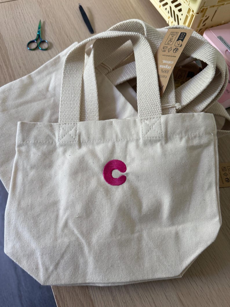 K&ouml;nnte beinhalten: Eine kleine, cremewei&szlig;e Canvas-Tragetasche mit zwei stabilen Henkeln und einem leuchtend pinkfarbenen, gestickten Buchstaben C in der Mitte. Ein braunes Papieretikett am Henkel tr&auml;gt Text, der unter anderem 100% Bio-Baumwolle angibt.