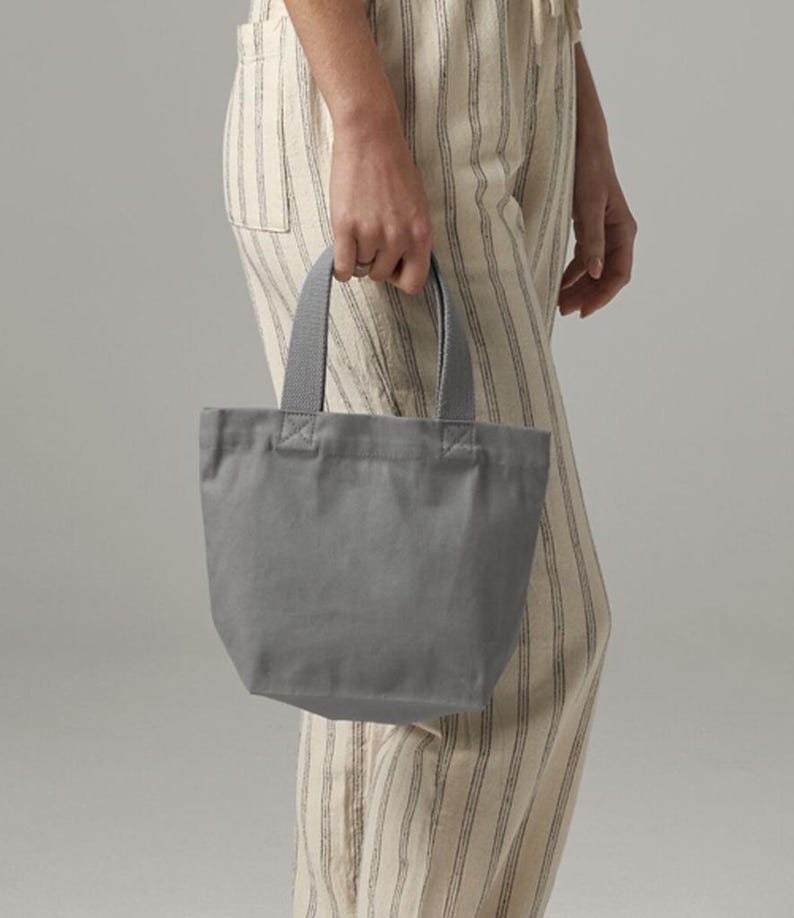 K&ouml;nnte beinhalten: Eine kleine, einfarbig graue Canvas-Tragetasche mit zwei stabilen Stoffgriffen, die mit verst&auml;rkten N&auml;hten befestigt sind. Die Tasche hat ein schlichtes Design mit offener Oberseite und einem strukturierten Boden f&uuml;r eine kompakte Aufbewahrung.