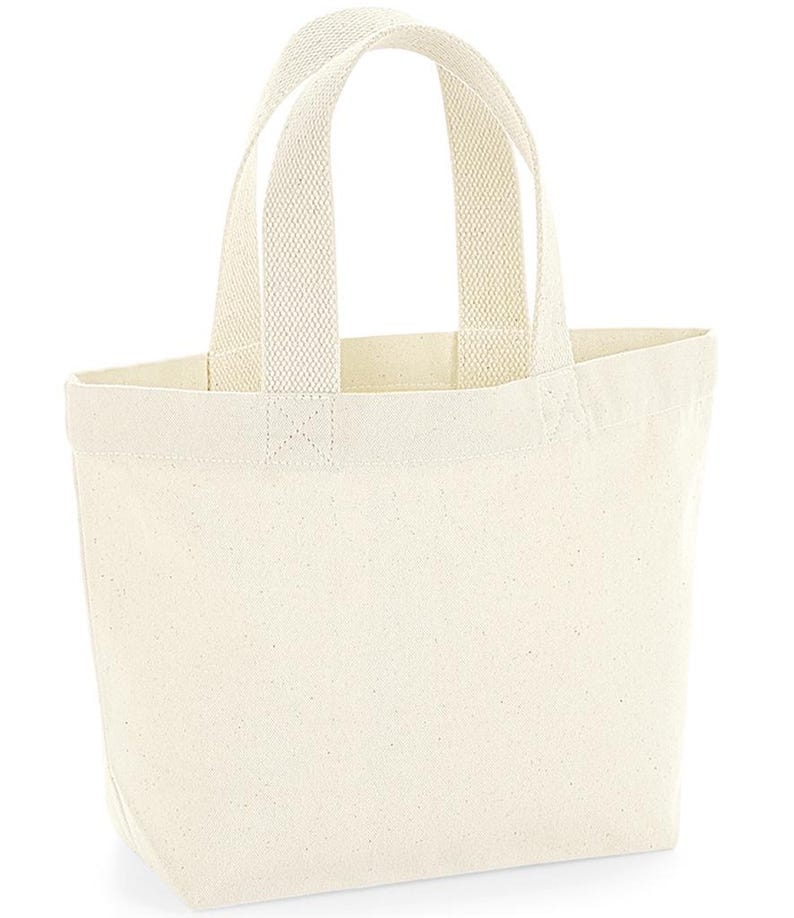 K&ouml;nnte beinhalten: Eine schlichte, cremewei&szlig;e Canvas-Einkaufstasche mit zwei stabilen, gewebten Tragegriffen an der Oberkante. Die Tasche hat eine einfache rechteckige Form mit flachem Boden und besteht aus einem strukturierten, naturfarbenen Stoff.