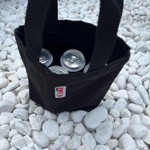 K&ouml;nnte beinhalten: Eine schwarze Tragetasche aus Segeltuch mit zwei stabilen Griffen, die mehrere silberne Getr&auml;nkedosen aus Aluminium enth&auml;lt. Ein kleines, wei&szlig;es, rechteckiges Stoffetikett mit dem roten Schriftzug Coke ist vorne mittig auf der Tasche vern&auml;ht.