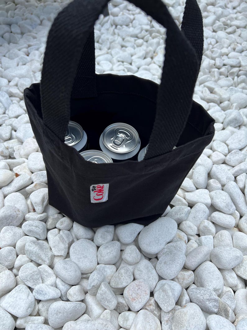 K&ouml;nnte beinhalten: Eine schwarze Tragetasche aus Segeltuch mit zwei stabilen Griffen, die mehrere silberne Getr&auml;nkedosen aus Aluminium enth&auml;lt. Ein kleines, wei&szlig;es, rechteckiges Stoffetikett mit dem roten Schriftzug Coke ist vorne mittig auf der Tasche vern&auml;ht.