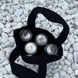 K&ouml;nnte beinhalten: Eine schwarze Stofftragetasche mit zwei stabilen Henkeln, die vier silberne Getr&auml;nkedosen aus Aluminium enth&auml;lt. Die Ansicht erfolgt direkt von oben und zeigt die kreisf&ouml;rmigen Dosen&ouml;ffner, die im dunklen Inneren der Tasche platziert sind.