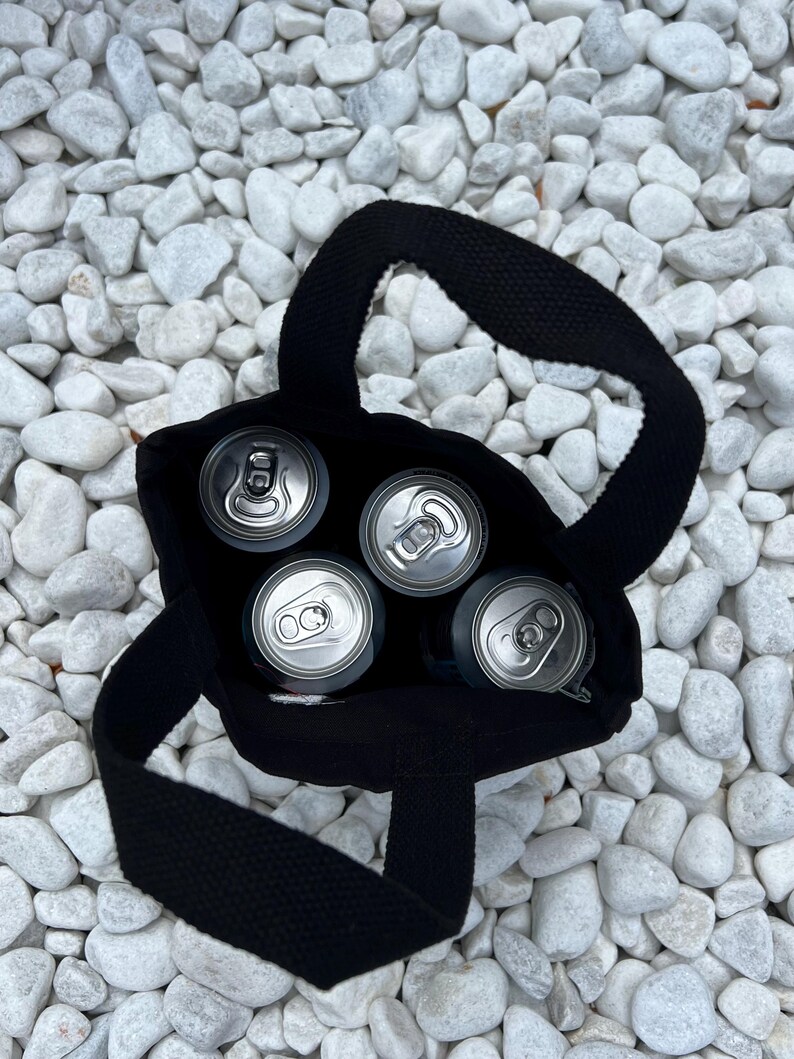K&ouml;nnte beinhalten: Eine schwarze Stofftragetasche mit zwei stabilen Henkeln, die vier silberne Getr&auml;nkedosen aus Aluminium enth&auml;lt. Die Ansicht erfolgt direkt von oben und zeigt die kreisf&ouml;rmigen Dosen&ouml;ffner, die im dunklen Inneren der Tasche platziert sind.