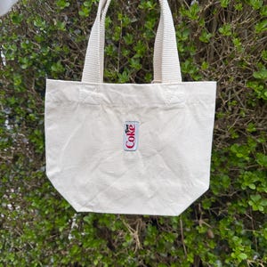 K&ouml;nnte beinhalten: Eine kleine, cremewei&szlig;e Canvas-Tragetasche mit zwei stabilen gewebten Griffen und einem rechteckigen Aufn&auml;her in der Mitte der Vorderseite. Der Aufn&auml;her zeigt das Diet Coke-Logo in rotem und schwarzem Text auf wei&szlig;em Grund mit einem d&uuml;nnen silbernen Rand.