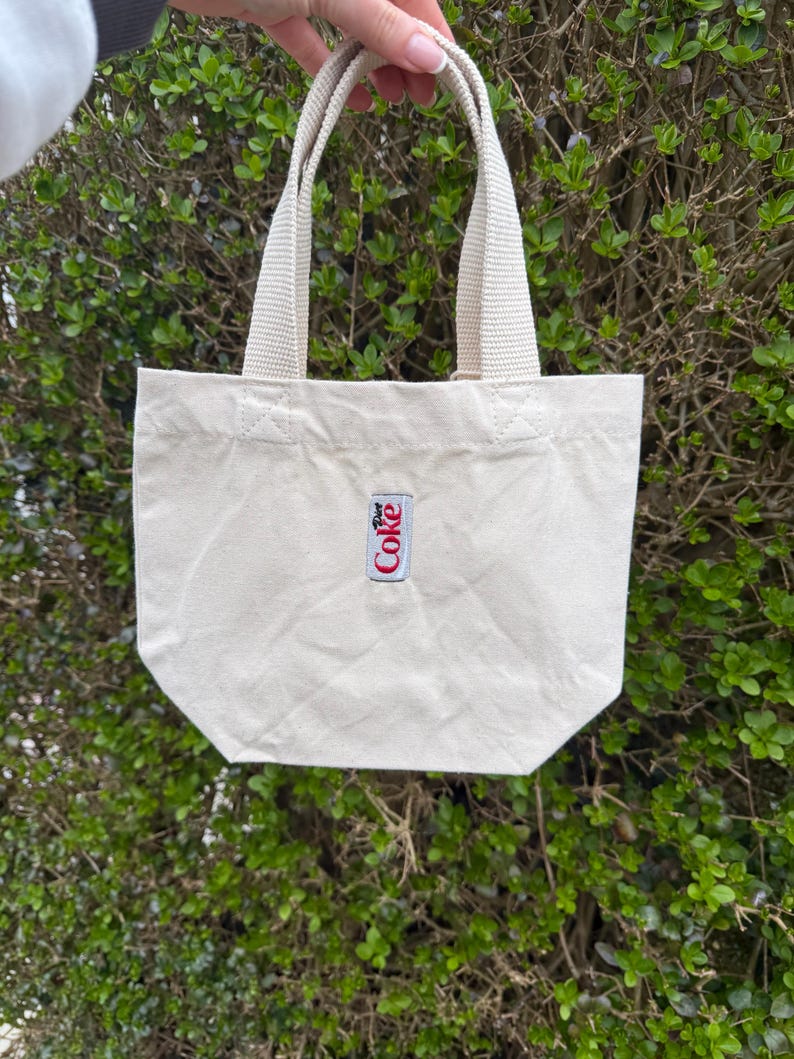 K&ouml;nnte beinhalten: Eine kleine, cremewei&szlig;e Canvas-Tragetasche mit zwei stabilen gewebten Griffen und einem rechteckigen Aufn&auml;her in der Mitte der Vorderseite. Der Aufn&auml;her zeigt das Diet Coke-Logo in rotem und schwarzem Text auf wei&szlig;em Grund mit einem d&uuml;nnen silbernen Rand.
