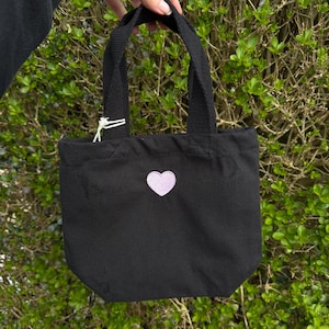 K&ouml;nnte beinhalten: Eine schwarze Tragetasche aus Segeltuch mit zwei stabilen schwarzen Stoffgriffen und einem kleinen, hellvioletten herzf&ouml;rmigen Aufn&auml;her in der Mitte der Vorderseite. Die Tasche hat ein schlichtes Design mit offener Oberseite und einem wei&szlig;en Etikett.