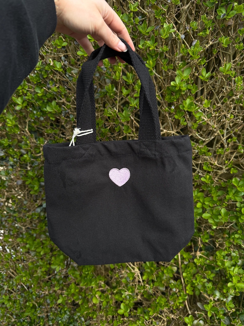 K&ouml;nnte beinhalten: Eine schwarze Tragetasche aus Segeltuch mit zwei stabilen schwarzen Stoffgriffen und einem kleinen, hellvioletten herzf&ouml;rmigen Aufn&auml;her in der Mitte der Vorderseite. Die Tasche hat ein schlichtes Design mit offener Oberseite und einem wei&szlig;en Etikett.