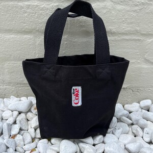 K&ouml;nnte beinhalten: Eine kleine schwarze Tragetasche aus Segeltuch mit zwei stabilen gewebten Griffen. Auf der Vorderseite befindet sich ein rechteckiger wei&szlig;er Aufn&auml;her mit dem roten, mittig aufgestickten Diet Coke-Logo. Die Tasche hat ein offenes Design.