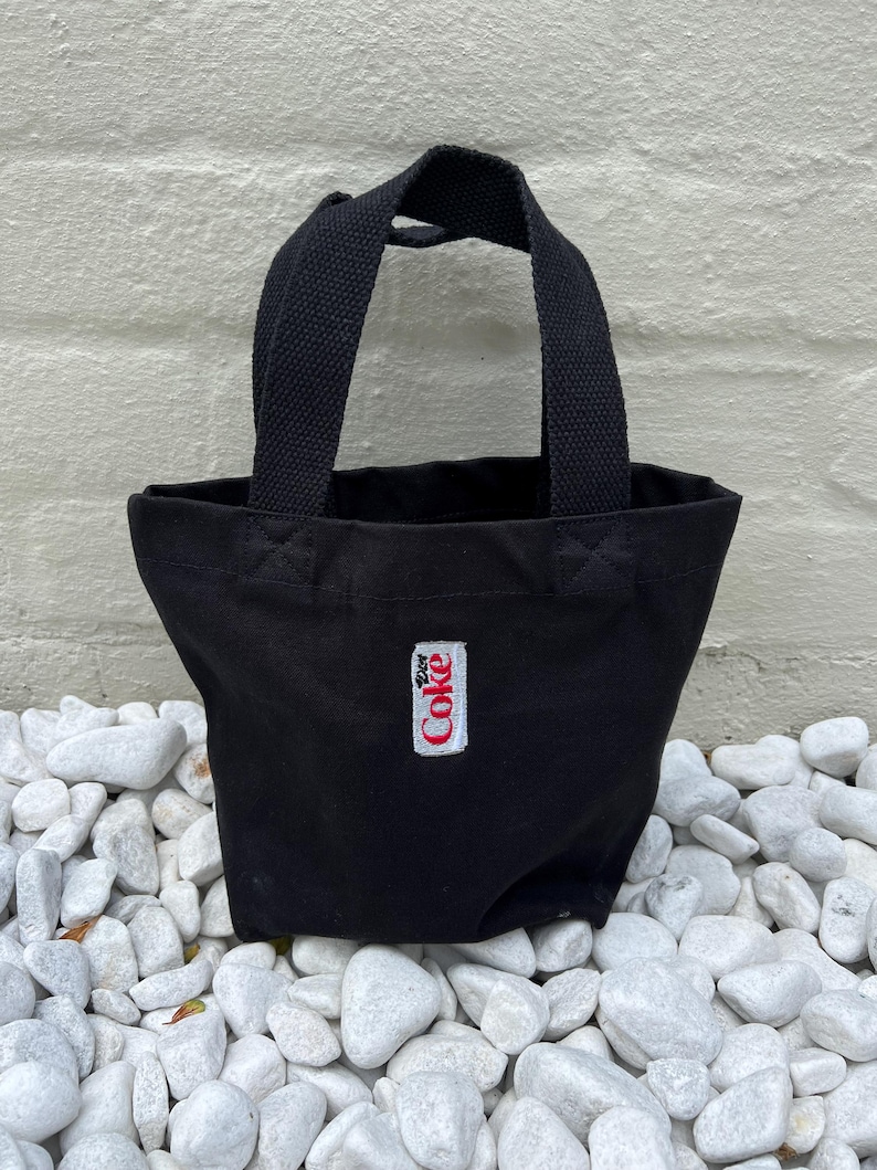 K&ouml;nnte beinhalten: Eine kleine schwarze Tragetasche aus Segeltuch mit zwei stabilen gewebten Griffen. Auf der Vorderseite befindet sich ein rechteckiger wei&szlig;er Aufn&auml;her mit dem roten, mittig aufgestickten Diet Coke-Logo. Die Tasche hat ein offenes Design.