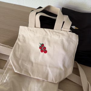 K&ouml;nnte beinhalten: Eine kleine, cremewei&szlig;e Canvas-Tragetasche mit einer aufgestickten Gruppe aus f&uuml;nf roten Tomaten mit gr&uuml;nen Stielen auf der Vorderseite. Die Tasche verf&uuml;gt &uuml;ber zwei robuste, farblich passende Stoffhenkel und ein einfaches, offenes Design.
