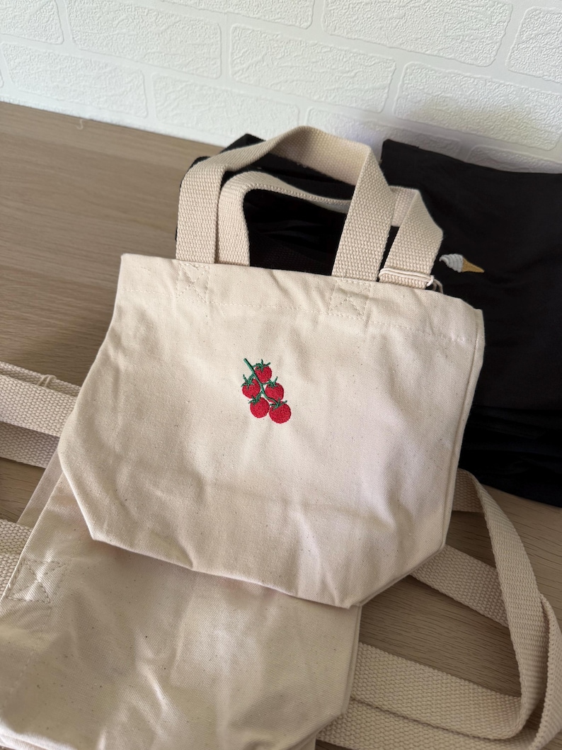 K&ouml;nnte beinhalten: Eine kleine, cremewei&szlig;e Canvas-Tragetasche mit einer aufgestickten Gruppe aus f&uuml;nf roten Tomaten mit gr&uuml;nen Stielen auf der Vorderseite. Die Tasche verf&uuml;gt &uuml;ber zwei robuste, farblich passende Stoffhenkel und ein einfaches, offenes Design.