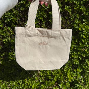 K&ouml;nnte beinhalten: Eine kleine, cremewei&szlig;e Canvas-Tragetasche mit zwei stabilen Tragegriffen und einem einzelnen, kleinen gestickten Croissant-Motiv in der Mitte der Vorderseite. Die Tasche besteht aus einem schlichten Webstoff mit sichtbaren N&auml;hten am Griffansatz.