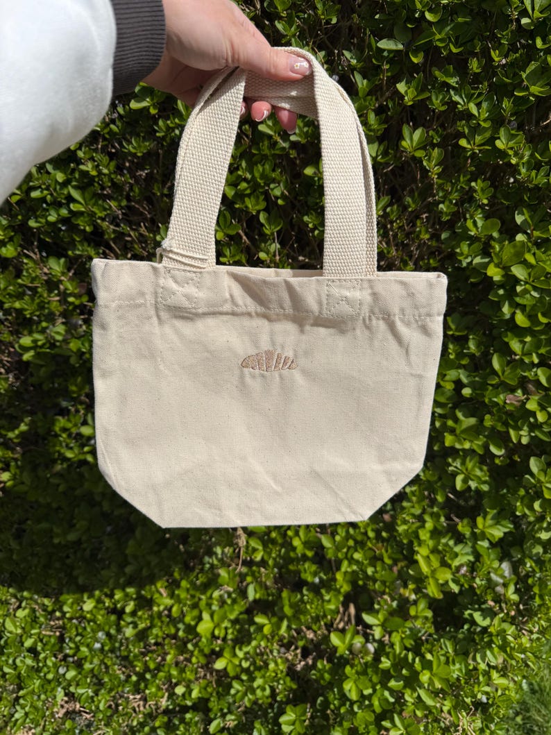 K&ouml;nnte beinhalten: Eine kleine, cremewei&szlig;e Canvas-Tragetasche mit zwei stabilen Tragegriffen und einem einzelnen, kleinen gestickten Croissant-Motiv in der Mitte der Vorderseite. Die Tasche besteht aus einem schlichten Webstoff mit sichtbaren N&auml;hten am Griffansatz.