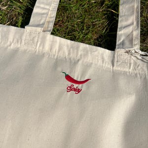 Könnte beinhalten: Cremefarbene Tragetasche mit zwei Henkeln. Die Tasche hat einen roten, gestickten Chili-Pfeffer mit dem Wort "Spicy" in roter Schreibschrift darunter. Die Tasche ist aus einem robusten Stoff gefertigt.
