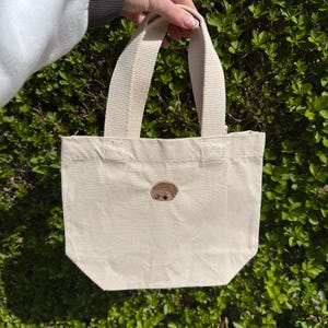 K&ouml;nnte beinhalten: Eine kleine, cremewei&szlig;e Canvas-Einkaufstasche mit einem mittig aufgestickten braunen Faultiergesicht. Die Tasche verf&uuml;gt &uuml;ber zwei robuste, gewebte Stoffgriffe an der Oberkante. Das Design ist schlicht, mit einem rechteckigen K&ouml;rper.