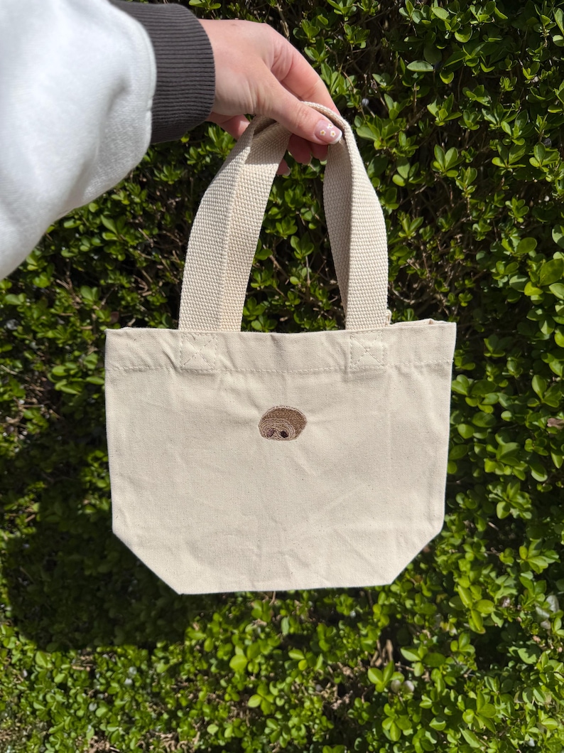 K&ouml;nnte beinhalten: Eine kleine, cremewei&szlig;e Canvas-Einkaufstasche mit einem mittig aufgestickten braunen Faultiergesicht. Die Tasche verf&uuml;gt &uuml;ber zwei robuste, gewebte Stoffgriffe an der Oberkante. Das Design ist schlicht, mit einem rechteckigen K&ouml;rper.