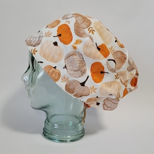 May include: A scrub cap featuring a white background with a repeating pattern of orange and beige pumpkins and small autumn leaves. The cap has a rounded, gathered shape with an adjustable elastic cord and toggle closure at the back for a secure fit.