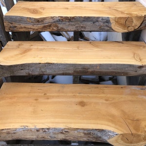 May include: Three wooden steps made from natural tree trunks. The steps are unfinished and have a rustic look. The steps are made from a light brown wood with a natural grain pattern.