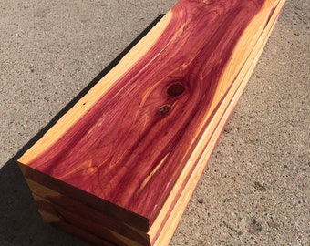 May include: Stacked planks of red cedar wood with a natural grain pattern. The wood has a reddish-purple hue and is smooth to the touch.