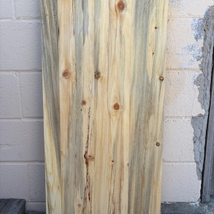 May include: A rectangular wooden plank with a natural, weathered appearance. The wood grain is prominently displayed, featuring a mix of light and dark tones, including beige, gray, and brown. Knots are visible throughout the plank, adding to its rustic charm. The text "*Wetted Down" is at the top.