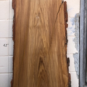 May include: A large, rectangular piece of wood with a live edge. The wood is a light brown color with a natural grain pattern. The wood is 42 inches tall, 25 inches wide, and 1.25 inches thick. The wood is labeled "ROAK 1".