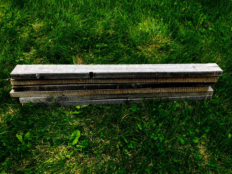 May include: A stack of weathered wooden boards, likely reclaimed wood, lying on a bed of green grass.