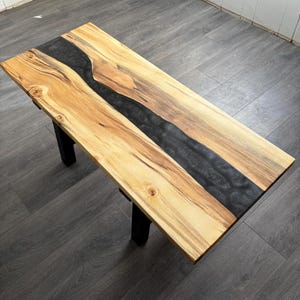 May include: A wooden coffee table with a black epoxy resin river running through the center. The table has black metal legs.
