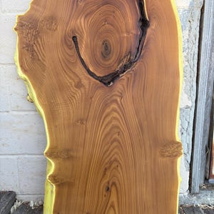 May include: A rectangular, live edge wooden slab with a natural, organic shape. The wood features a warm, brown color with prominent grain patterns and a dark, knot-like feature. The edges are a lighter yellow-green color.