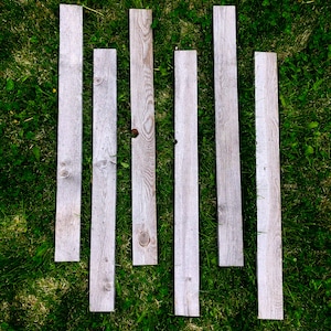 May include: Six weathered wooden planks laid out on a bed of green grass. The planks are all the same width and length, but vary slightly in color and texture.