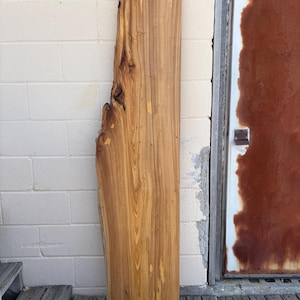 May include: A rectangular, unfinished wooden slab with a natural edge. The wood has a warm, golden-brown color with prominent grain patterns and knots. The slab is leaning against a white brick wall and a weathered door.