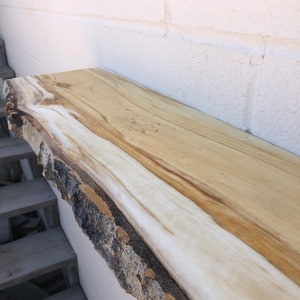 May include: A natural wood shelf with a live edge and a light brown and white wood grain. The shelf is mounted to a white brick wall.