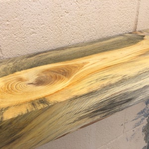 May include: A close-up of a wooden shelf with a natural wood grain pattern. The wood has a blue and yellow color variation. The shelf is mounted on a white brick wall.