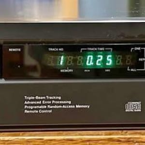 Könnte beinhalten: Schwarzer ADC CD-Player mit digitaler Anzeige von Titel und Zeit. Funktionen umfassen Wiedergabe/Pause, Stopp/Öffnen und Titelsteuerung. Das Gerät hat einen Power-Knopf und ein CD-Fach.