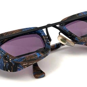 Puede incluir: Gafas de sol con un diseño único. Las monturas negras presentan un patrón azul y marrón. Las lentes son moradas. Las gafas de sol tienen una forma moderna y angular.