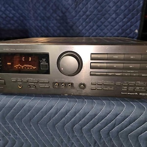Puede incluir: Un receptor de procesador acústico digital JVC RX-888V plateado. El panel frontal presenta una gran perilla de volumen, numerosos botones y una pantalla que muestra texto iluminado en naranja. El receptor tiene múltiples puertos de entrada y salida.
