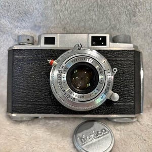Puede incluir: Una cámara Konica Konirapid-S vintage con una lente Hexanon 1:2.8. El cuerpo de la cámara es negro con detalles plateados y un anillo de lente plateado. Una tapa de lente plateada con el logotipo de Konica está debajo.