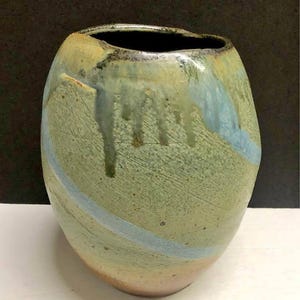 Puede incluir: Un jarrón de cerámica hecho a mano con una forma orgánica única. El jarrón presenta una superficie texturizada con una mezcla de tonos verdes, azules y beige. Gotas de esmalte añaden interés visual, y una franja azul lo rodea. La abertura es oscura.
