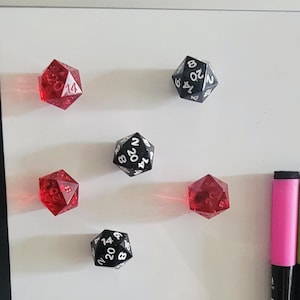 May include: A white dry-erase board with several red and black polyhedral dice and colorful dry-erase markers. The dice are numbered and have various faces. The markers are pink, green, and blue.