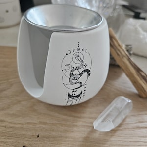 Könnte beinhalten: Weiß-grauer Diffusor für ätherische Öle mit einem schwarzen Schlangen- und Monddesign. Ein klarer Quarzkristall und ein Holzstab befinden sich neben dem Diffusor. Der Diffusor hat einen silbernen Rand und ein geschwungenes Design.