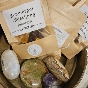 Könnte beinhalten: Eine Sammlung von Natursteinen und kleinen Papiertüten in einer messingfarbenen Schale. Die Tüten haben Etiketten mit deutschem Text. Zu den Steinen gehören ein weißer Mondstein, ein grün-roter Stein, ein brauner Stein und ein violetter Amethyst.
