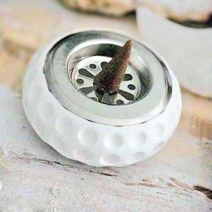 Könnte beinhalten: Ein weißer Keramik-Räucherstäbchenhalter mit einer genoppten Oberfläche, die einem Golfball ähnelt. Er hat einen silbernen Metalleinsatz mit floralem Muster und hält einen braunen Räucherkegel. Der Halter steht auf einer hellen Oberfläche.