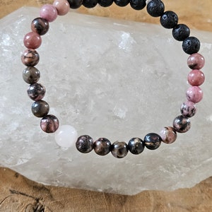 Könnte beinhalten: Ein Perlenarmband mit rosa, braunen und schwarzen Perlen. Das Armband hat eine weiße Perle und wird auf einer hellen Oberfläche präsentiert. Die Perlen sind wahrscheinlich aus verschiedenen Steinarten gefertigt.