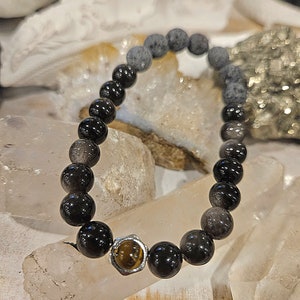 Könnte beinhalten: Ein Armband mit schwarzen und grauen kugelförmigen Perlen. Das Armband hat eine zentrale Perle mit einer goldfarbenen Fassung und einem Tigeraugenstein. Das Armband wird auf einer hellen Oberfläche präsentiert.