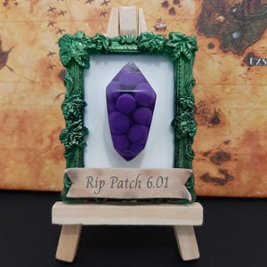May include: A small framed picture with a green frame and a purple crystal inside. The frame is on a small wooden easel. The text on the frame reads "Rip Patch 6.01".