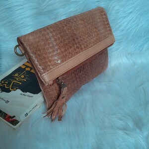 May include: A woven leather clutch in shades of tan and light brown, featuring a fold-over flap and a zipper closure with a tassel. The clutch has a ring on the side and is resting on a white, fluffy surface.