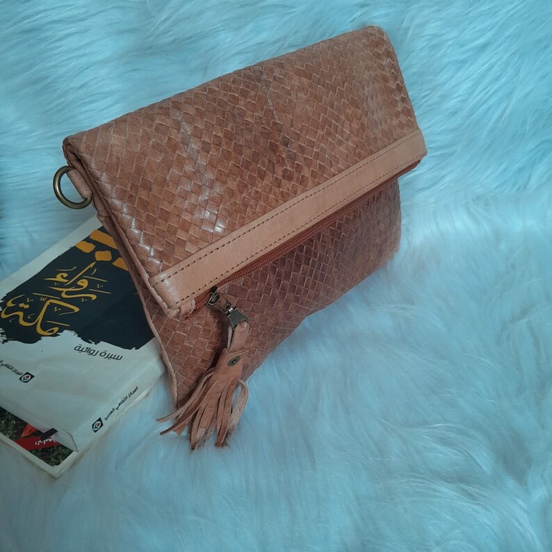 May include: A woven leather clutch in shades of tan and light brown, featuring a fold-over flap and a zipper closure with a tassel. The clutch has a ring on the side and is resting on a white, fluffy surface.