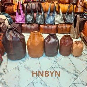 Könnte beinhalten: Eine Auswahl an Lederhandtaschen und -taschen in verschiedenen Stilen und Farben. Das Bild zeigt Kordelzug-Eimertaschen in Braun- und Beigetönen sowie Umhängetaschen und Satteltaschen. Die Taschen werden auf einem marmorierten Boden präsentiert.